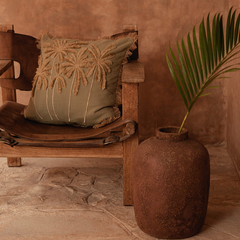 Wooden chair with a decorative pillow against a textured wall, next to a rustic vase and plant.